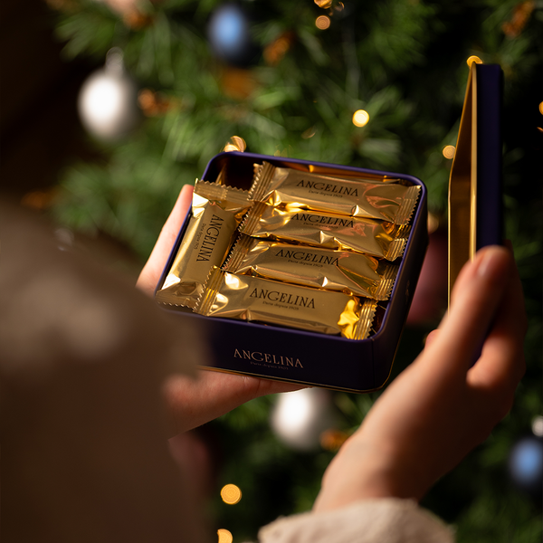 A person holds an elegant tin box of Angelina's crispy crepes wrapped in golden foil, with a Christmas tree decorated with lights and ornaments blurred in the background.