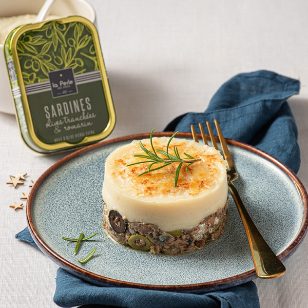 tin of sardines with sliced olives and rosemary with a recipe made with them on a plate in the foreground