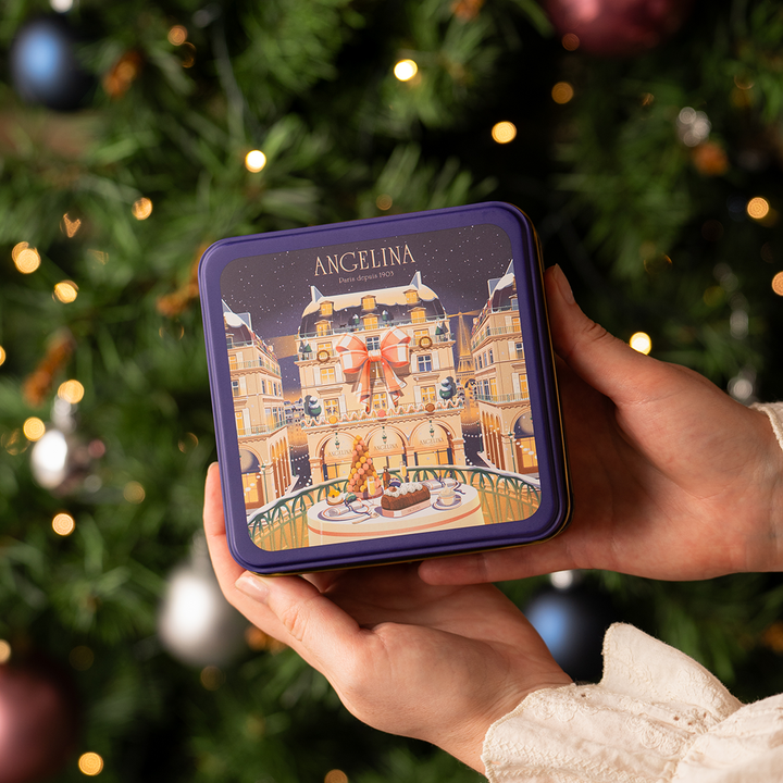 A person holds a festive Angelina Paris collector tin with an illustrated winter café scene on the lid, in front of a decorated Christmas tree with twinkling lights and ornaments.