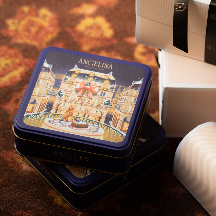A festive Angelina Paris collector tin with an illustrated winter café scene on the lid sits stacked on a patterned carpet beside elegant white gift boxes tied with black ribbons.