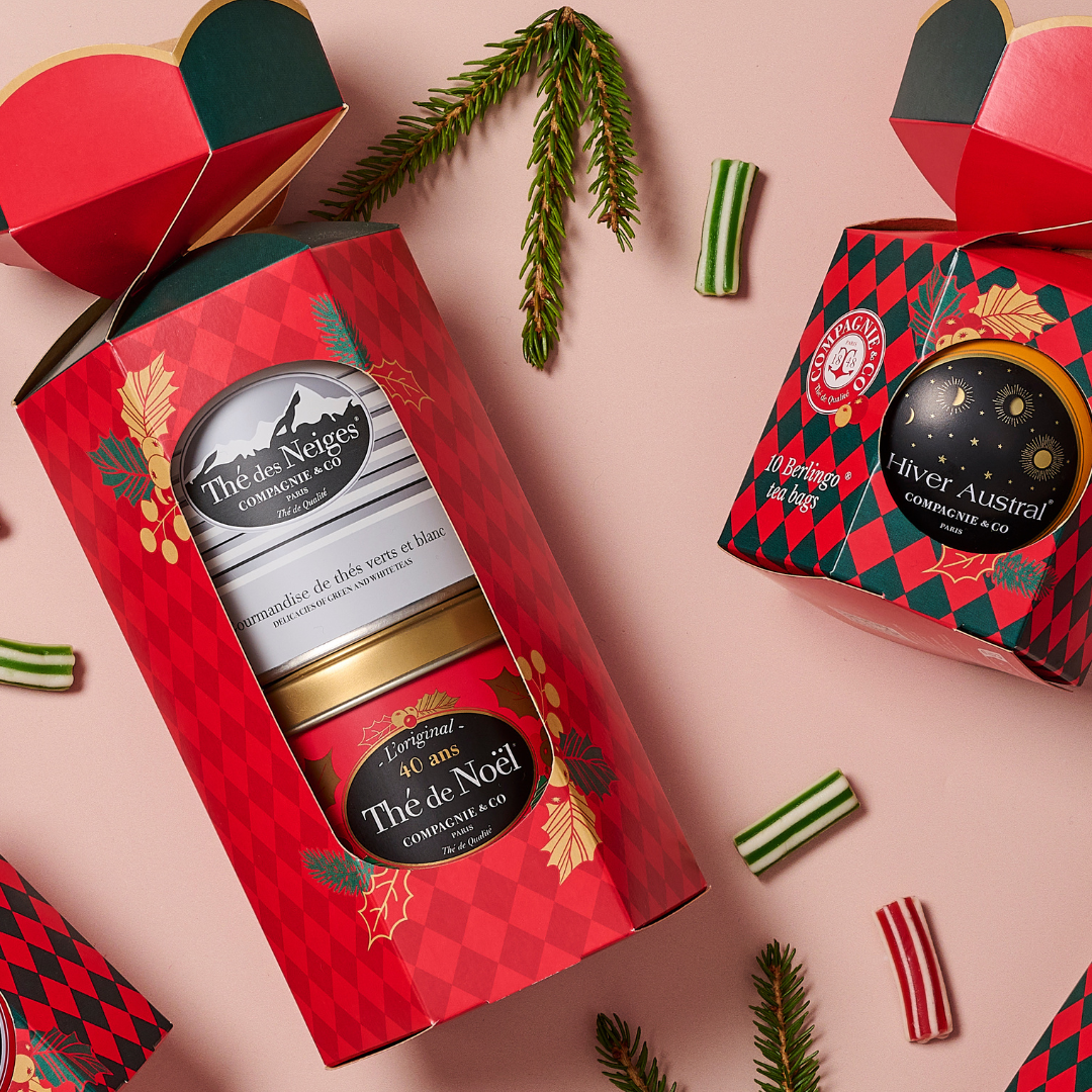 Festive Compagnie &amp; Co tea gift sets displayed on a pink surface, featuring decorative red and green packaging with tins labeled “Thé des Neiges” and “Thé de Noël,” surrounded by candy pieces and pine sprigs.
