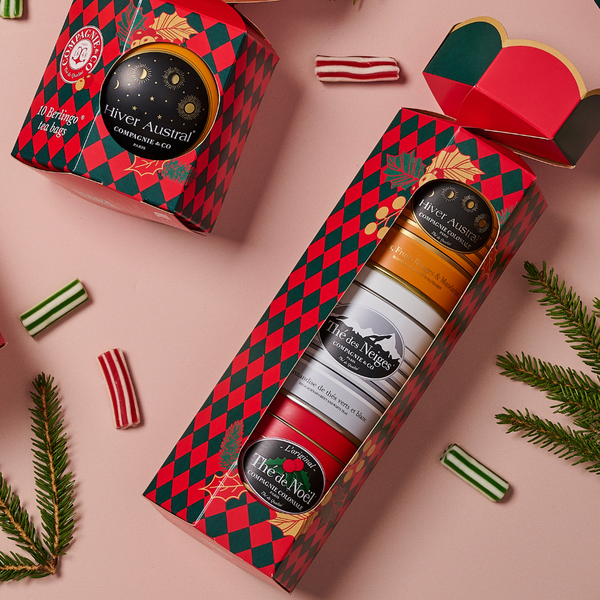Compagnie & Co holiday tea gift sets arranged on a pink surface, featuring festive red and green diamond-patterned boxes with tins labeled “Hiver Austral,” “Thé des Neiges,” and “Thé de Noël,” surrounded by striped candies and pine branches.